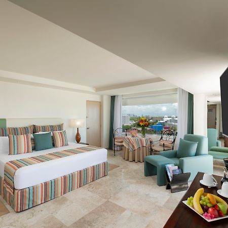 Grand Oasis Palm Hotel Cancún Exterior foto The photo shows a spacious hotel room with modern decor. It features a large bed with a striped duvet and decorative pillows, flanked by two lamps. There are comfortable seating areas with light green chairs and a small table. A balcony can be seen i