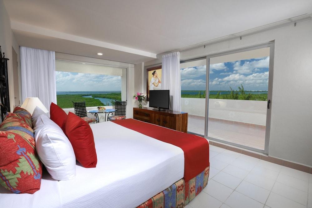 Grand Oasis Palm Hotel Cancún Exterior foto The photo shows a modern hotel room featuring a large bed with white linens and red accents. There are decorative pillows on the bed, and a bedside table with a lamp. To the side, there’s a large window with white curtains that offers a view of a bal