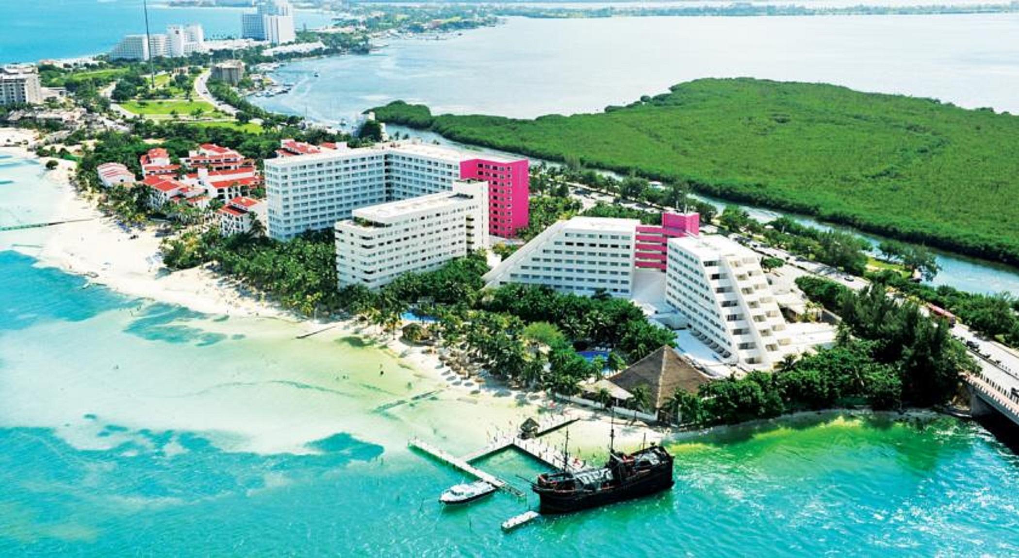 Grand Oasis Palm Hotel Cancún Exterior foto The photo shows a coastal resort area featuring several large hotel buildings, some of which are white and others are pink. The hotels are situated near a sandy beach, with clear blue water stretching out into the background. There are palm trees and