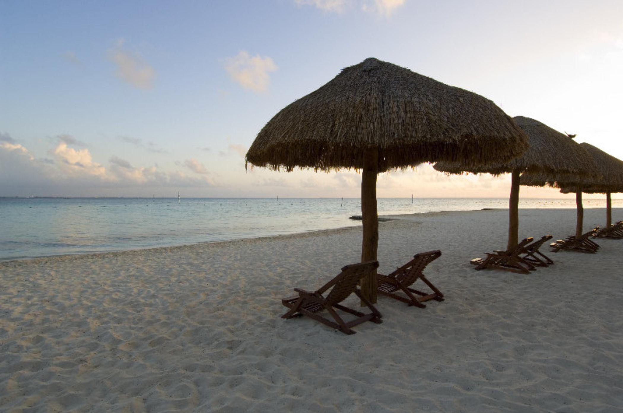 Grand Oasis Palm Hotel Cancún Exterior foto The photo depicts a serene beach scene featuring several thatched umbrellas on the sand. Beneath the umbrellas are wooden lounge chairs, positioned for relaxation. The beach has soft, white sand, and the calm waters of the sea reflect the colors of t