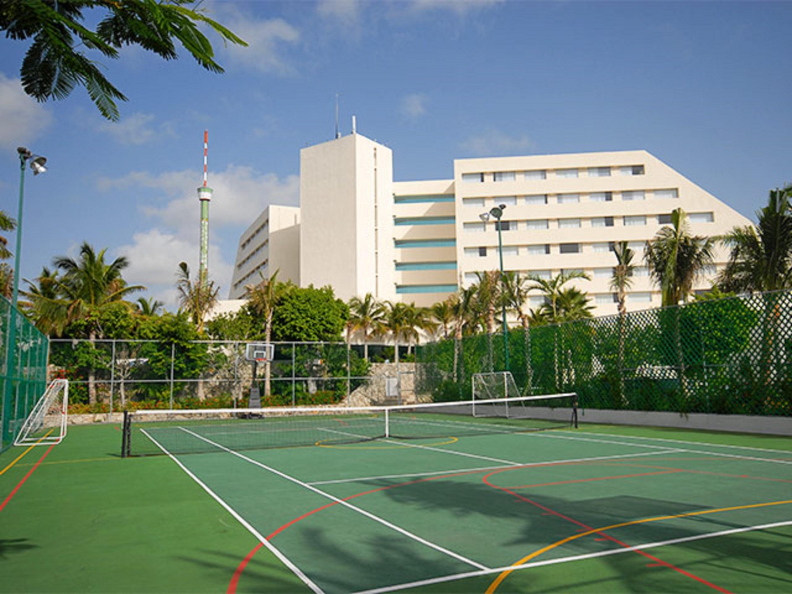 Grand Oasis Palm Hotel Cancún Exterior foto The photo shows a sports facility featuring a tennis court in the foreground. The court is surrounded by a green fence and has distinctive lines marking the playing area. In the background, there is a tall, modern building, possibly a hotel or resort