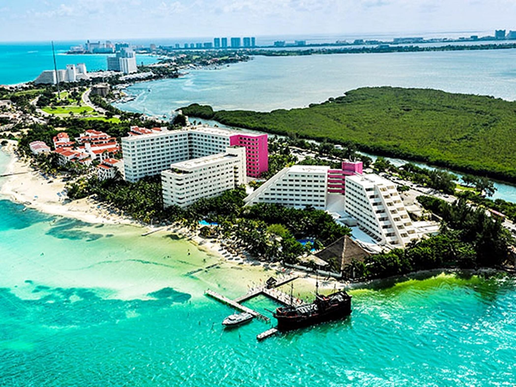 Grand Oasis Palm Hotel Cancún Exterior foto The photo depicts an aerial view of a coastal area, showcasing several buildings along the shoreline. The structures include a mix of white and pink hotels or resorts, surrounded by lush greenery and sandy beaches. The water is a vibrant turquoise co