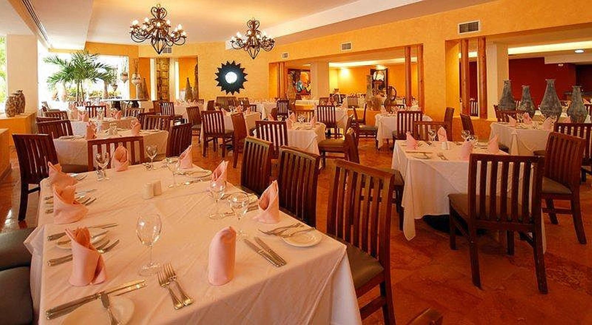 Grand Oasis Palm Hotel Cancún Restaurant foto The photo shows an interior of a restaurant with an inviting and elegant ambiance. There are several tables set up, each covered with white tablecloths. The place settings include glassware, silverware, and neatly folded pink napkins. The walls are p