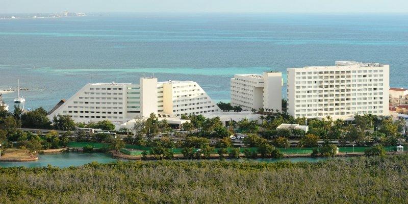 Grand Oasis Palm Hotel Cancún Exterior foto The photo shows a coastal scene featuring several large hotel buildings. These structures are modern, with a mix of white walls and geometric shapes. They are situated near the ocean, which has clear turquoise waters. In the foreground, there is gree